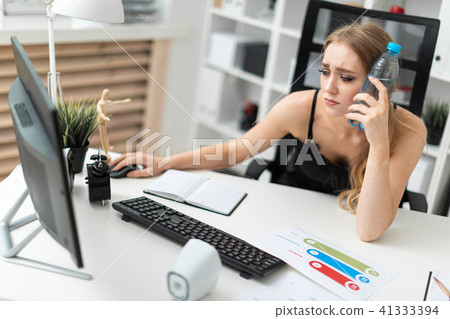 A young girl sits at a computer desk in the office and puts a bottle of water to her head. A young girl sits at a computer desk in the office and puts a bottle of water to her head. 41333394