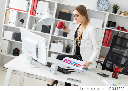 The young girl sat down on the table in her office, holding a red cup in her hand and working with a The young girl sat down on the table in her office, holding a red cup in her hand and working with a 41333531