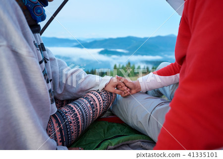 Close-up of guy and girl holding hands sitting in tent on blurred background mountains Close-up of guy and girl holding hands sitting in tent on blurred background mountains 41335101