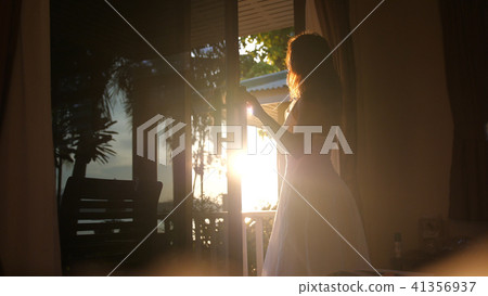 Beautiful brunette girl stands in the doorway in the early morning during sunrise and looks to the 41356937