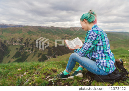 Portrait of a young hipster girl with multi-colored hair sitting on nature in the mountains reading 41372422