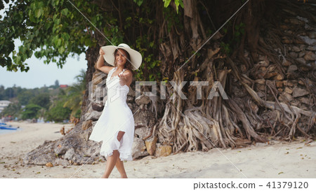 Young brunette woman with long hair in white drees and hat beckons to follow her on tropical beach 41379120
