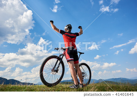 Athletic sportsman cyclist standing with bike on top of hill, rasing hands, against blue sky Athletic sportsman cyclist standing with bike on top of hill, rasing hands, against blue sky 41401645