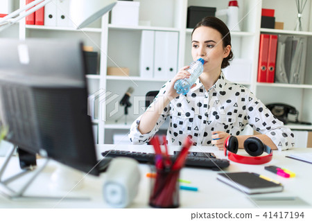A beautiful young girl sits at a table in the office and drinks water from a bottle. 41417194