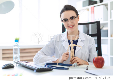 A beautiful young girl in a white robe is sitting at a table and holding a wooden figure of a man in 41417196