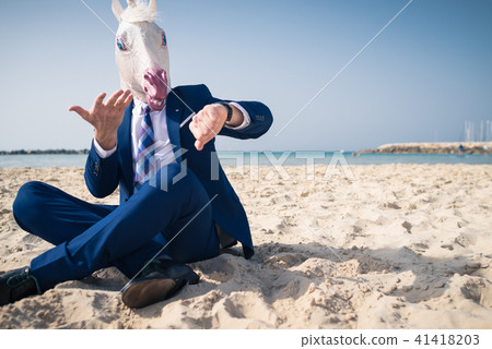 Stylish man in funny mask and elegant suit sits on beach 41418203