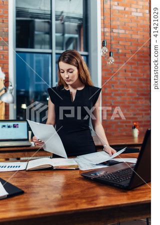 Serious woman reading papers studying resumes standing at work desk in stylish office Serious woman reading papers studying resumes standing at work desk in stylish office 41421029