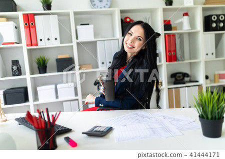 A beautiful young girl is holding a glass with coffee, sitting on a chair in the office at the table 41444171