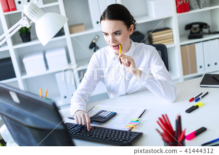 A young girl in the office holds a pen in her mouth and works with a calculator, documents and a 41444312