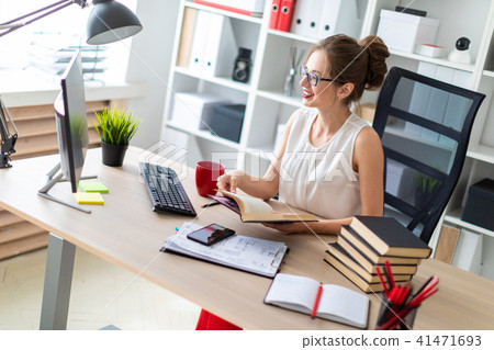 A young girl sits at a computer desk and holds an open book in her hands and looks at the monitor. A young girl sits at a computer desk and holds an open book in her hands and looks at the monitor. 41471693