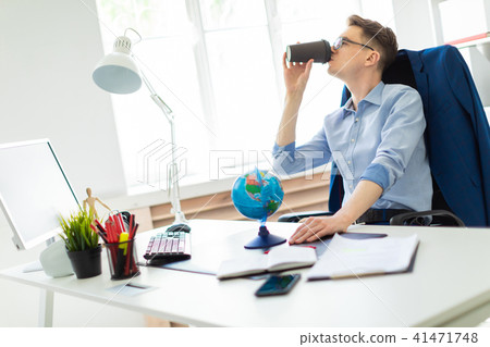 A young man sits in the office at a computer desk, holds a globe with his hand and drinks coffee. 41471748