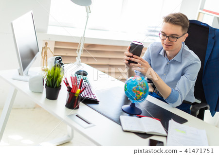 A young man sits in the office at a computer desk and holds a glass of coffee in his hand. A young 41471751