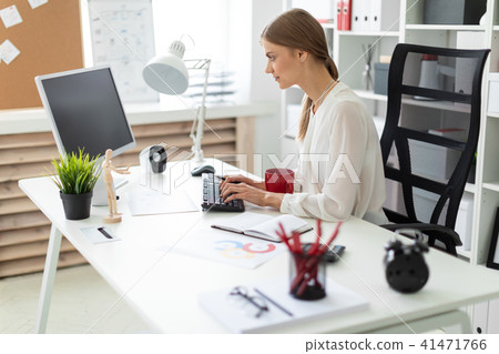 A young girl is sitting at a table in the office, holding a red cup in her hand and working at the A young girl is sitting at a table in the office, holding a red cup in her hand and working at the 41471766