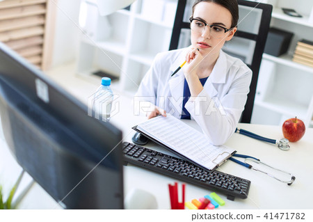 A beautiful young girl in a white robe is sitting at a computer desk with documents and a pen in her 41471782