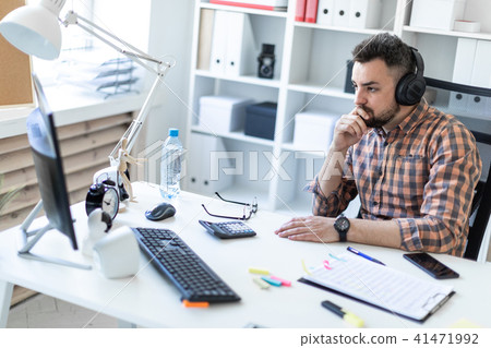 A young man in headphones sits at a table in the office and looks at the monitor. A young man in headphones sits at a table in the office and looks at the monitor. 41471992