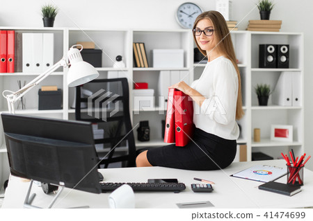 The young girl sat down on the table in the office and holds a folder of documents. The young girl sat down on the table in the office and holds a folder of documents. 41474699