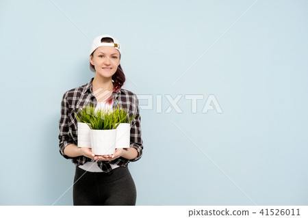 A girl in work clothes and a white cap is holding pots with green indoor plants against the blue A girl in work clothes and a white cap is holding pots with green indoor plants against the blue 41526011