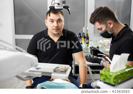 Male dentist in a black uniform is looking to the camera while another is working with dental 41563083