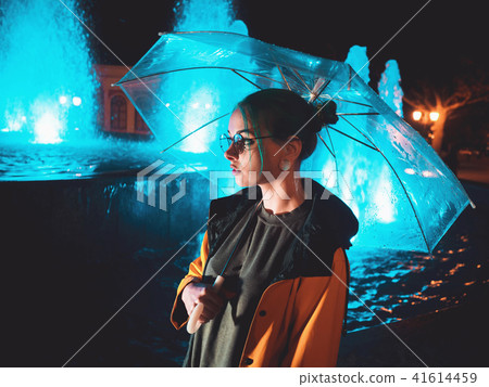 Young pretty girl with blue dyed hair in yellow raincoat and with transparent umbrella stands near 41614459
