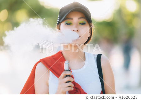Pretty young hipster asian woman in black hat vape ecig, vaping device at the sunset. Toned image Pretty young hipster asian woman in black hat vape ecig, vaping device at the sunset. Toned image 41620525