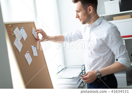 A young man stands near a board with stickers and holds a pen in his hand. A young man tears off a 41634531