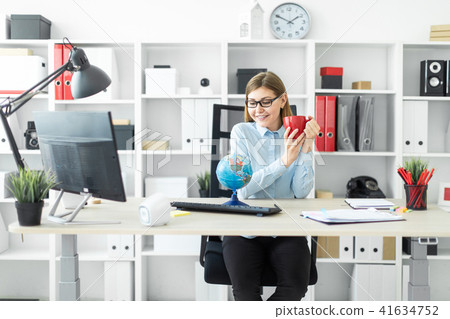 A young girl in glasses is sitting at a table, holding a red cup in her hands and looking at the A young girl in glasses is sitting at a table, holding a red cup in her hands and looking at the 41634752