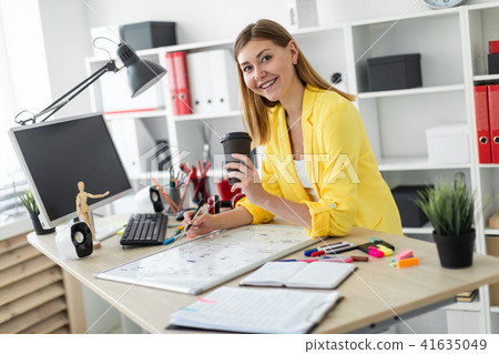 A young girl stands near a table and holds a glass of coffee and a marker. Before the girl on the 41635049