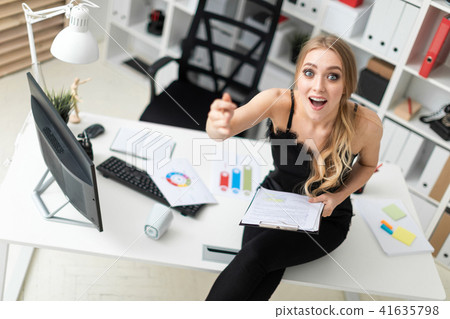 A young girl sits on a table in the office and holds a pen and a tablet with sheets. 41635798