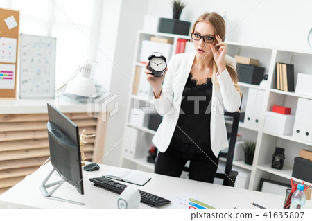 A young girl stands near a table in the office and holds an alarm clock. A young girl stands near a table in the office and holds an alarm clock. 41635807