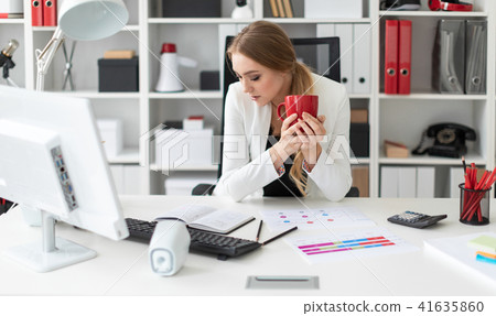 A young girl sits at a computer desk in the office and holds a red cup in her hand. Before the girl A young girl sits at a computer desk in the office and holds a red cup in her hand. Before the girl 41635860