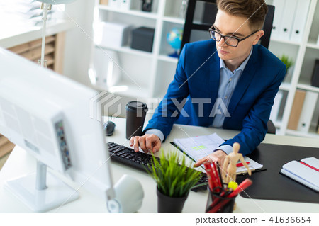 A young man sits at a table in the office and works with documents and a computer. A young man sits at a table in the office and works with documents and a computer. 41636654
