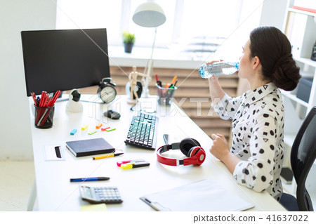 A beautiful young girl sits at a table in the office and drinks water from a bottle. 41637022