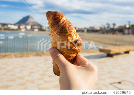 Croissant in hand against the backdrop of a beautiful beach Croissant in hand against the backdrop of a beautiful beach 41659643
