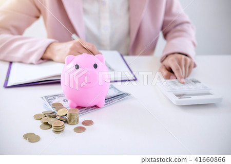 Business woman putting coins into piggy bank, writing to report Business woman putting coins into piggy bank, writing to report 41660866