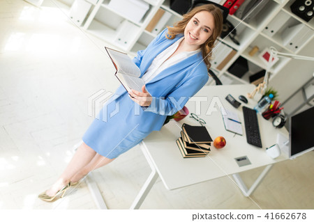 A beautiful young girl stands near a table in the office and holds an open book in her hands. A beautiful young girl stands near a table in the office and holds an open book in her hands. 41662678