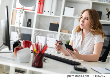 Beautiful young girl sits in the office, holds a bank card and phone in her hand. Beautiful young girl sits in the office, holds a bank card and phone in her hand. 41662679