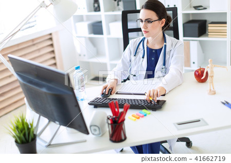 A beautiful young girl in a white robe is sitting at a computer desk with documents and a pen in her 41662719