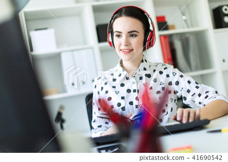Beautiful young girl sits in headphones at desk in office, holds a notebook in her hand and prints 41690542