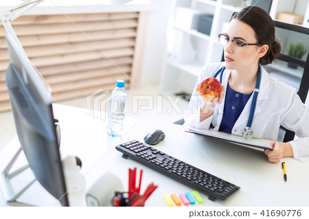 A beautiful young girl in a white robe is sitting at a computer desk with documents and an apple in 41690776