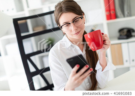 A young girl is sitting at a table in the office, holding a phone and a red cup in her hand. 41691083