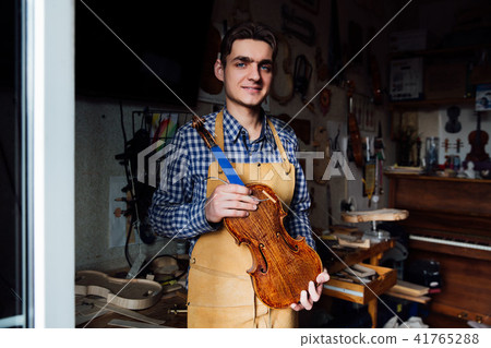 portrait of a young luthier with a handcrafted violin in his workshop with the tools. 41765288