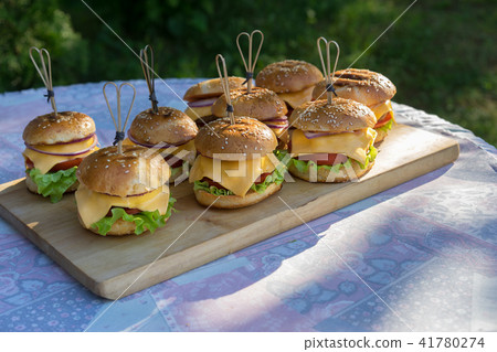 Tasty burgers with cheese, lettuce, onion and tomatoes served outdoor on a wooden table. Tasty burgers with cheese, lettuce, onion and tomatoes served outdoor on a wooden table. 41780274