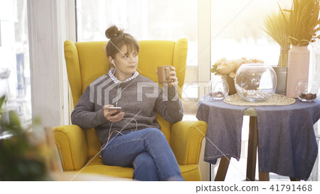 A young girl sitting in a big chair with a phone listening to music drinking tea A young girl sitting in a big chair with a phone listening to music drinking tea 41791468