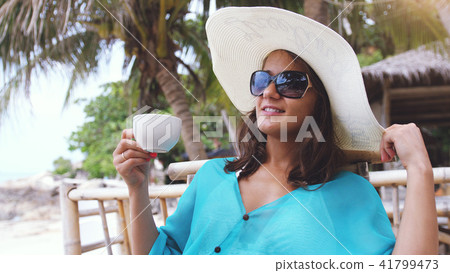 Elegant pretty girl in hat and sunglasses on holiday, sitting in beach cafe with scenic views of the Elegant pretty girl in hat and sunglasses on holiday, sitting in beach cafe with scenic views of the 41799473