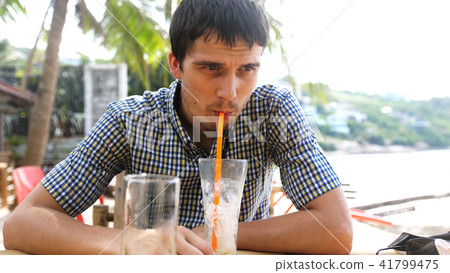 Handsome young man in beautifu shirt drinking cocktail in beach cafe sitting by sea Handsome young man in beautifu shirt drinking cocktail in beach cafe sitting by sea 41799475