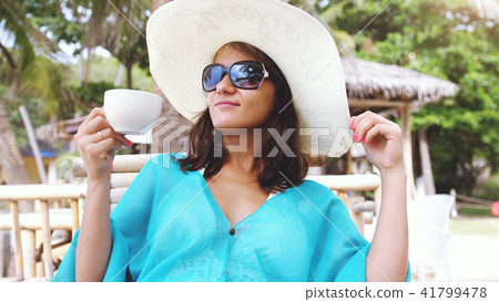 Young elegant pretty woman wearing hat and sunglasses on holiday, sitting in beach cafe drinks Young elegant pretty woman wearing hat and sunglasses on holiday, sitting in beach cafe drinks 41799478
