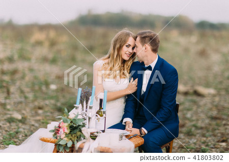 Close-up horizontal view of the hugging newlyweds sitting at the wedding table on the coast. 41803802