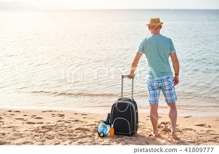Man tourist in summer clothes with a suitcase in his hand, looking at the sea on the beach, concept 41809777