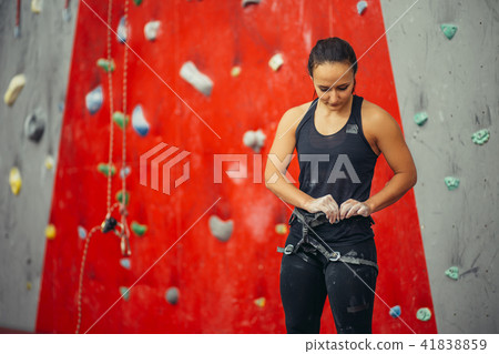 Climber woman puts on belaying harness for practice on artificial rock wall. Climber woman puts on belaying harness for practice on artificial rock wall. 41838859