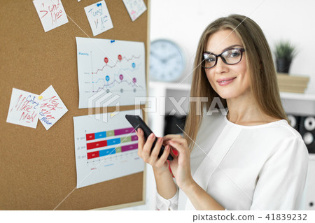 A young girl stands near a board with stickers and holds a phone in her hands. A young girl stands near a board with stickers and holds a phone in her hands. 41839232